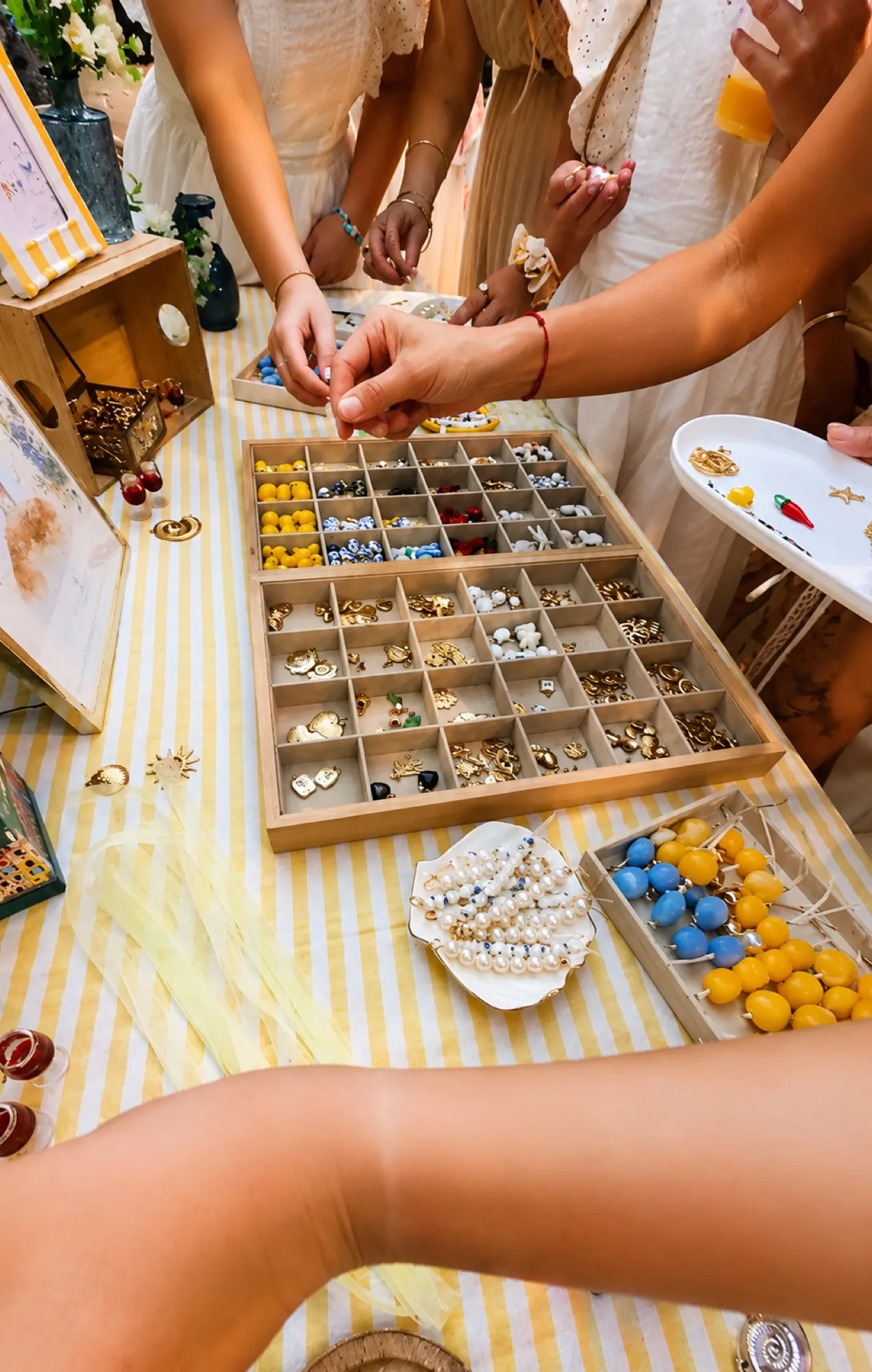 Invitées composant leur bijou autour du plateau de charms, sur une nappe rayée jaune