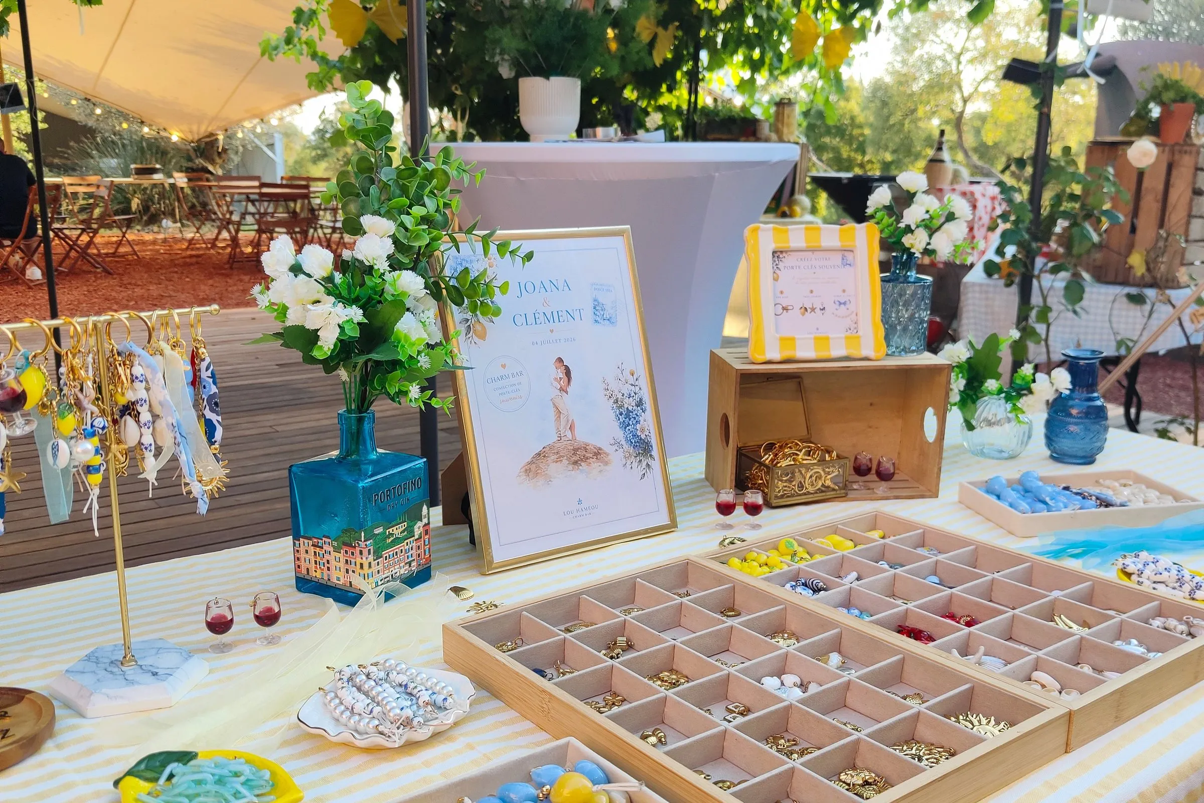 Le Charm Bar dressé sous la tonnelle : nappe rayée, panneau Joana & Clément, fleurs blanches et plateaux de charms